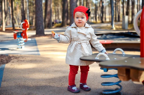 Doce menina pequena loira criança em casaco de trincheira bege bonita e boné vermelho joga ao ar livre no parque infantil — Fotografia de Stock