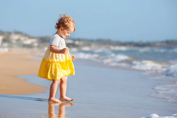 Lindo bebé rizado niña con un vestido amarillo jugando en una hermosa playa tropical —  Fotos de Stock