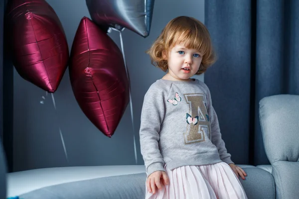 Retrato de menina feliz com cabelo loiro encaracolado em roupas casuais posando dentro de casa com balões estrela no fundo — Fotografia de Stock