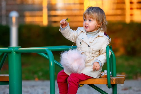 Porträtt av Söt blond liten baby flicka i trenchcoat och fuskpäls väska på lekplats — Stockfoto