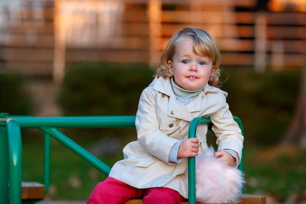 Retrato de doce loira pequena menina em casaco de trincheira e saco de pele falsa no parque infantil — Fotografia de Stock