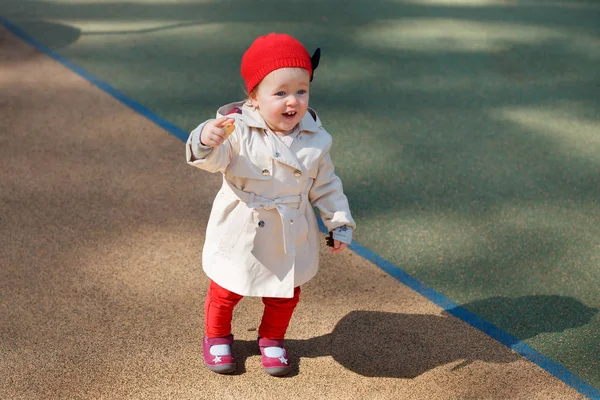 Doce menina pequena loira criança em casaco de trincheira bege bonita e boné vermelho joga ao ar livre no parque infantil — Fotografia de Stock