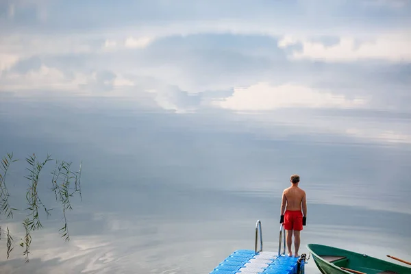 若い男は湖の桟橋に単独で立ちます。村の湖の日の出 ストック写真