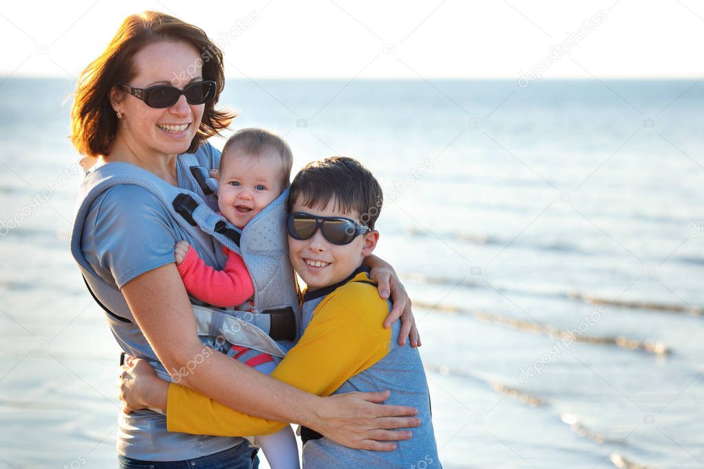 Young mother with her two children on sea at summer day. Big brother and adorable baby girl in a carrier backpack