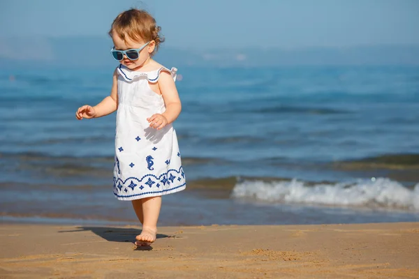Niedliche lockige Baby-Mädchen spielen an einem schönen tropischen Strand trägt ein weißes Kleid lizenzfreie Stockfotos