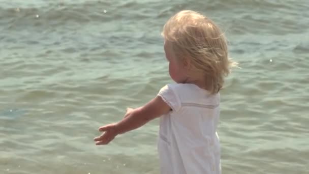 Vackra lilla tjej går längs havsstranden. Liten flicka i en vit klänning på kusten. Slow motion. — Stockvideo