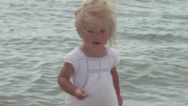Güzel küçük bir kız deniz kıyısı yürür. Deniz kenarı beyaz elbiseli küçük kız. Ağır çekim. — Stok video
