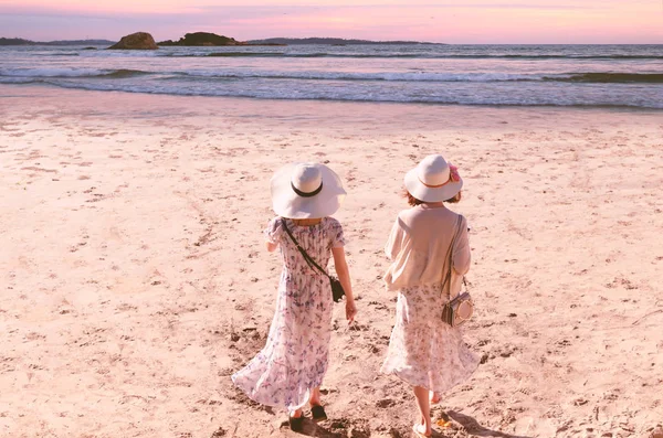 Two young friend  women in long dresses and hats walk along the beach to sea at evening sunset time. The concept of feminism, independence of women of friendship. Toned in instagram style.