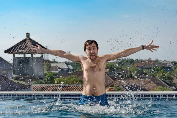 Portrait of joyful slender man with chest hair, emerges from pool, water splashes, shows tongue and hands up, on hotel roof. Emotions - joy, happiness, excited, positive, cheery, gleeful, glad.