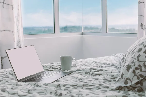 Morning in modern apartment - open laptop is standing on bed in front of large window, next to cup of coffee, side view. Beginning of the day. Concept work at home, always always in touch, connected.