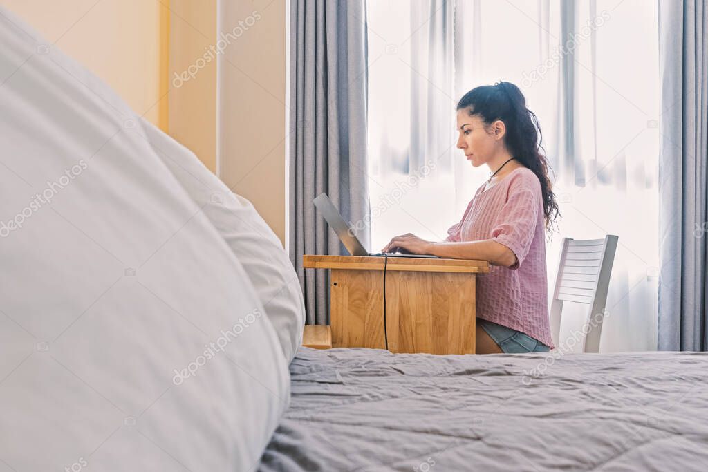 Portrait of a serious beautiful young woman working on a laptop at home. In the foreground is a bed with pillows. Photo with selective soft focus.