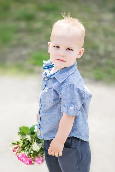 Portrait of small baby boy standing with beautiful roses bouquet outdoor at sunner park. Little gentleman with flowers looking into camera at summer sunny day — Stock Photo, Image