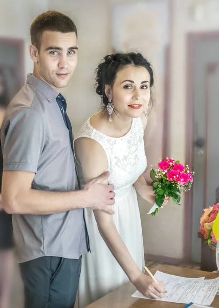 Cérémonie officielle d'enregistrement du mariage au bureau d'enregistrement en Ukraine. Jeune beau couple mariée et marié mettre des signatures sur la forme. Mariée souriante tient stylo et petit bouquet de mariage . — Photo