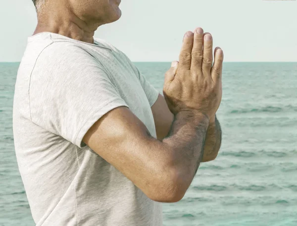 An adult man stands by joining his palms in front of him in a gesture namaste by the sea.