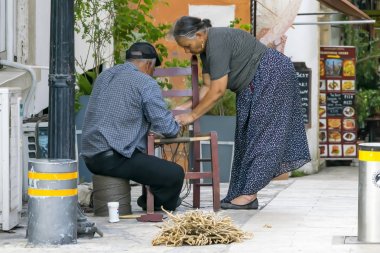 CYPRUS, NICOSIA - JUNE 10, 2019: Elderly Greek couple of craftsmen making wicker chairs on a city street. Family traditional business. clipart
