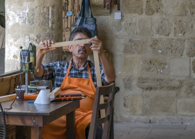 Kıbrıs, Lefkoşa - 10 Haziran 2019: Yerel bir sanat galerisinde oturan ve çalışan yaşlı bir zanaatkar portresi. İşyerinde bir elektrikli makine üzerinde çalışan doğrama