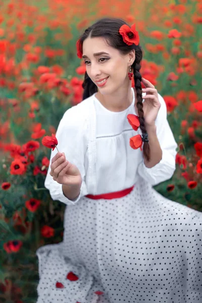 Porträtt av en vacker ung kvinna i bakgrunden av ett blommande vallmofält. Leende ukrainska kvinna sitter och håller en vallmo blomma — Stockfoto