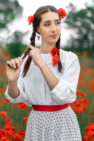 Young pretty girl Slavic or Ukrainian posing in folk dress on a flowering poppy field. Female weaving braid. — Stock Photo, Image