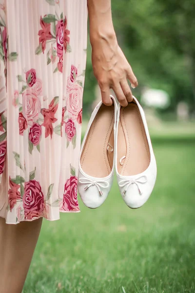 Молодая красивая женщина в летнем платье ходит по лугу по зеленой траве. Молодая девушка держит в руках белые балетные туфли. — стоковое фото