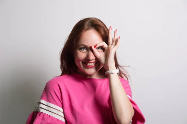 Närbild porträtt av vackra glada rödhårig kaukasiska kvinnliga leende, visar vita tänder, tittar på kameran genom fingrarna i okej gest. — Stockfoto