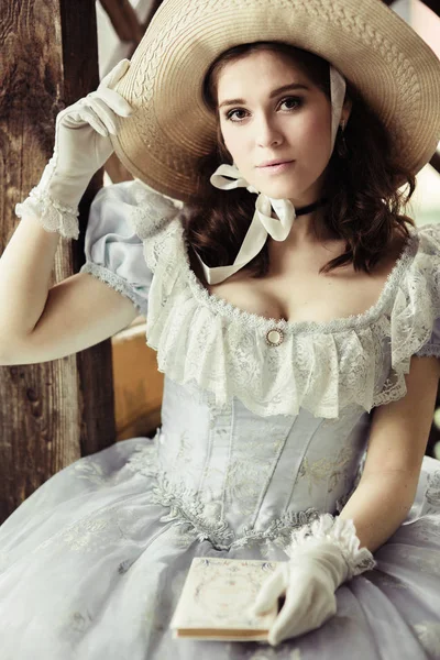 beautiful girl in a hat, historical dress, gloves, with a book in her hands sitting in the gazebo