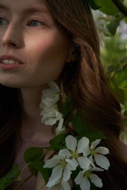 Sarı saçlı genç bir kızın yüzünün bir parçası, Elma ağaçlarının yapraklarının arasında dikiliyor ve başka tarafa bakıyor.                               