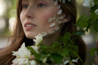 Açık renk saçlı genç bir kızın portresi, Elma ağaçlarının yapraklarının arasında duruyor ve yanaklarında beyaz elma yapraklarına bakıyor.                                 