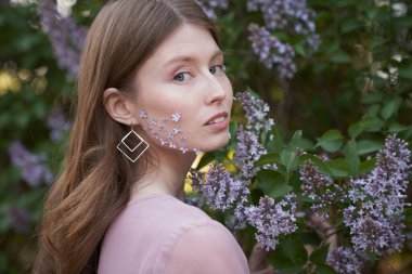 Açık renk uzun dalgalı saçları ve mavi gözleri olan genç bir kız, kızın yanaklarındaki leylak ağaçlarının yapraklarında dikilirken, doğrudan kameraya bakıyor. Kulaklarda geometrik şekiller var.                                