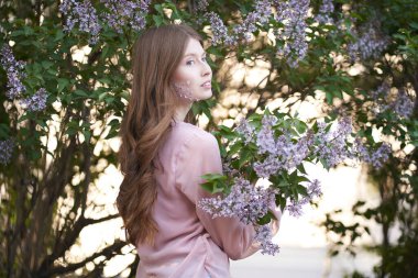 Açık renk uzun dalgalı saçları ve mavi gözleri olan genç bir kız, kızın yanaklarındaki ağaçların yapraklarında dikiliyor, uzaklara bakıyor.                               