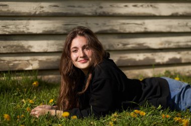 Siyah ceketli esmer genç bir kız yeşil çimlerin üzerinde uzanıp doğrudan kameraya bakıyor. Ahşap kalasların arkasında                                 