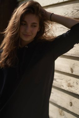   Uzun dalgalı saçlı, genç bir esmer kız, ahşap bir evin yanında duruyor, saçlarını eliyle tutuyor ve başını çeviriyor.                                  