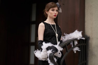 Kırmızı dudaklı, siyah retro elbiseli, şapkalı güzel esmer kadın. Doğrudan kameraya bakıyor, ahşap bir kapının arkasında.                               