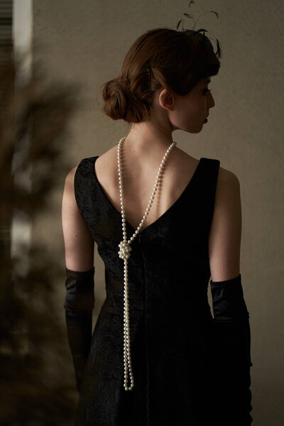 A beautiful brunette in a black retro hat stands with her back to the camera and looks away. Historical reconstruction                               