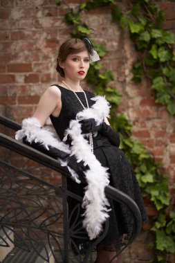 Kırmızı dudaklı, siyah retro elbiseli, şapkalı güzel esmer kadın. Tuğladan bir duvarın önünde dikilip uzaklara bakıyorsun.                               