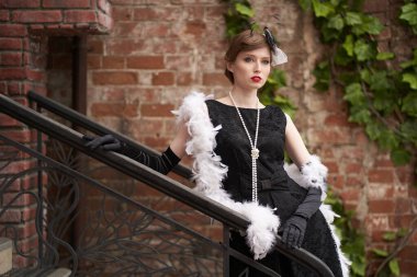 Kırmızı dudaklı, siyah retro elbiseli, şapkalı güzel esmer kadın. Tuğladan bir duvarın önünde dikilip uzaklara bakıyorsun.                               