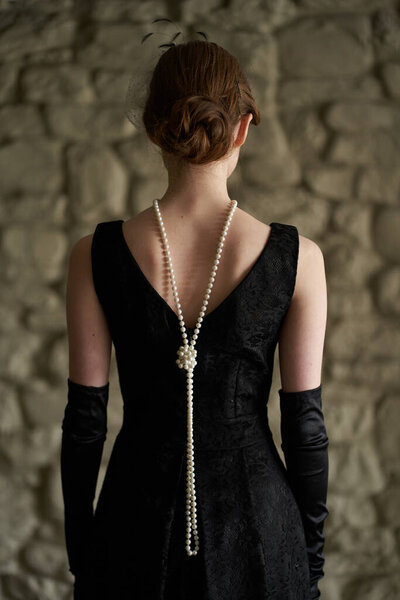 A beautiful brunette in a black retro hat stands with her back to the camera. Historical reconstruction                               