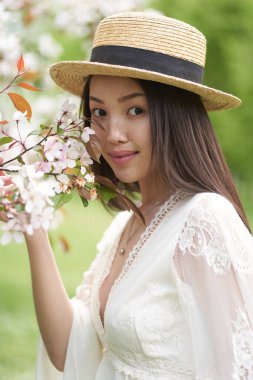 Beyaz elbiseli güzel bir Asyalı kadın, beyaz çiçekli bir ağacın yanında duruyor ve elinde dallarla kameraya bakıyor.                               