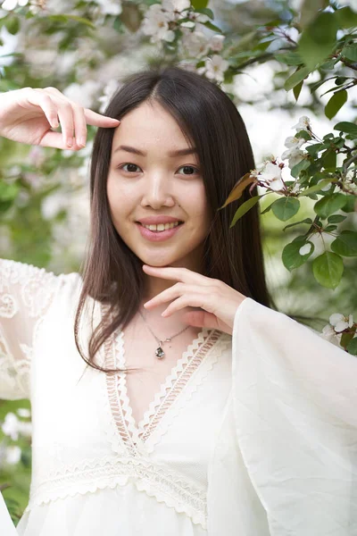 Beyaz elbiseli güzel bir Asyalı kadının portresi, beyaz çiçekli bir ağacın yanında duruyor, ellerini yüzüne yaklaştırıyor ve doğrudan kameraya bakıyor.                               