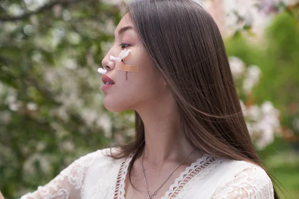 Beyaz elbiseli güzel bir Asyalı kadın, elinde beyaz çiçeklerle bir ağacın yanında duruyor, başka tarafa bakıyor.                               