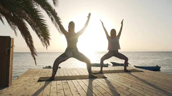 Silueta de dos mujeres practican diosa postura de yoga a orillas del mar bajo el sol —  Fotos de Stock