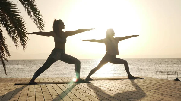 Silueta de dos mujeres practican postura guerrera de yoga a orillas del mar bajo el sol —  Fotos de Stock