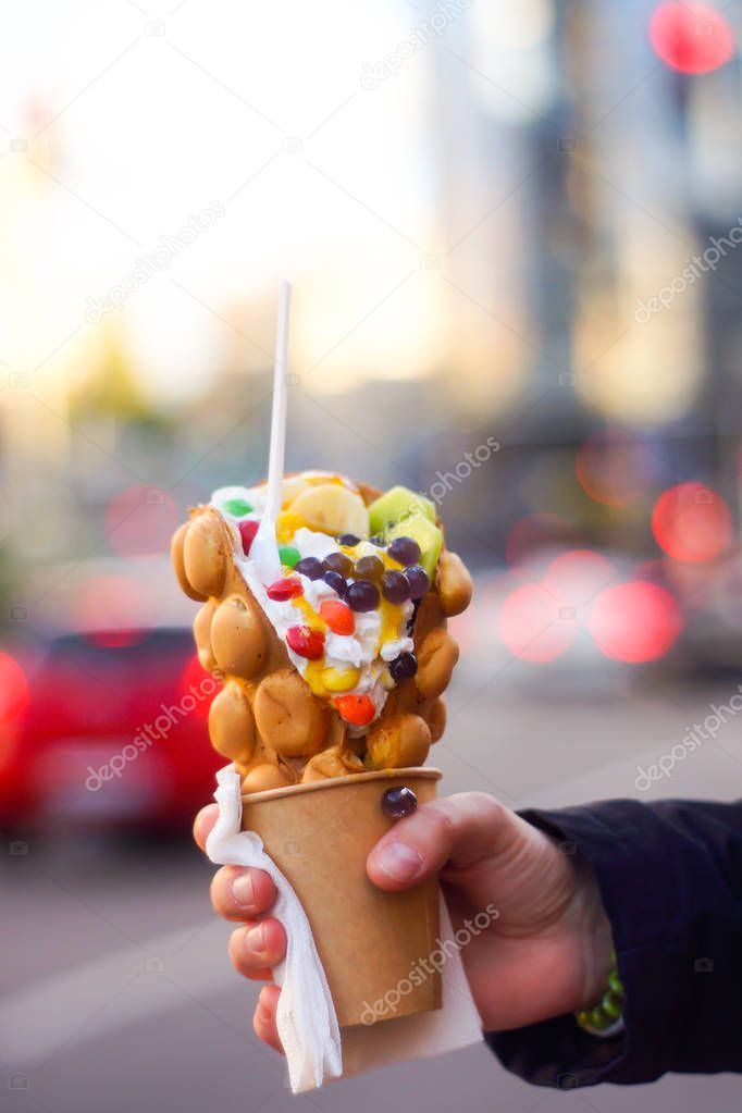 Hand holding bubble waffle with fruits, chocolate and marshmallow, with copy space.
