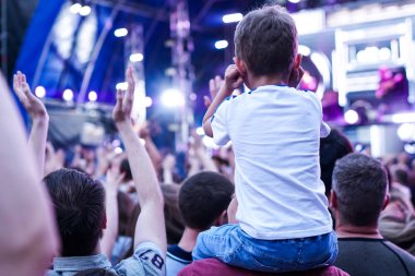 Çocuk bir açık hava rock müzik konserinde onlarla el ele tutuşarak ailesinin omuzlarında eğlenceli
