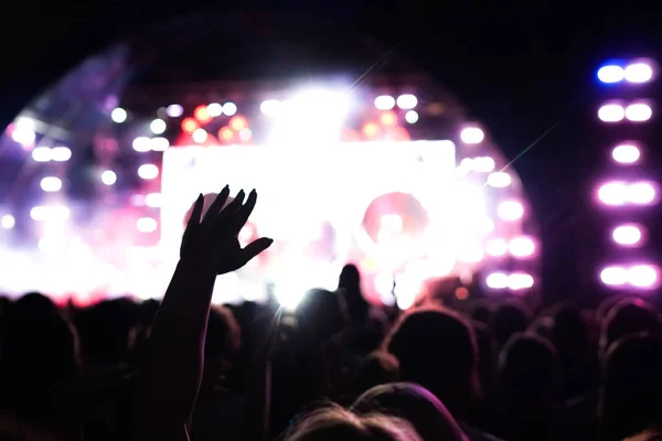 Multitud de conciertos asistiendo a un concierto, las siluetas de la gente son visibles, retroiluminadas por luces de escenario. Las manos levantadas y los teléfonos inteligentes son visibles aquí y allá . — Foto de Stock