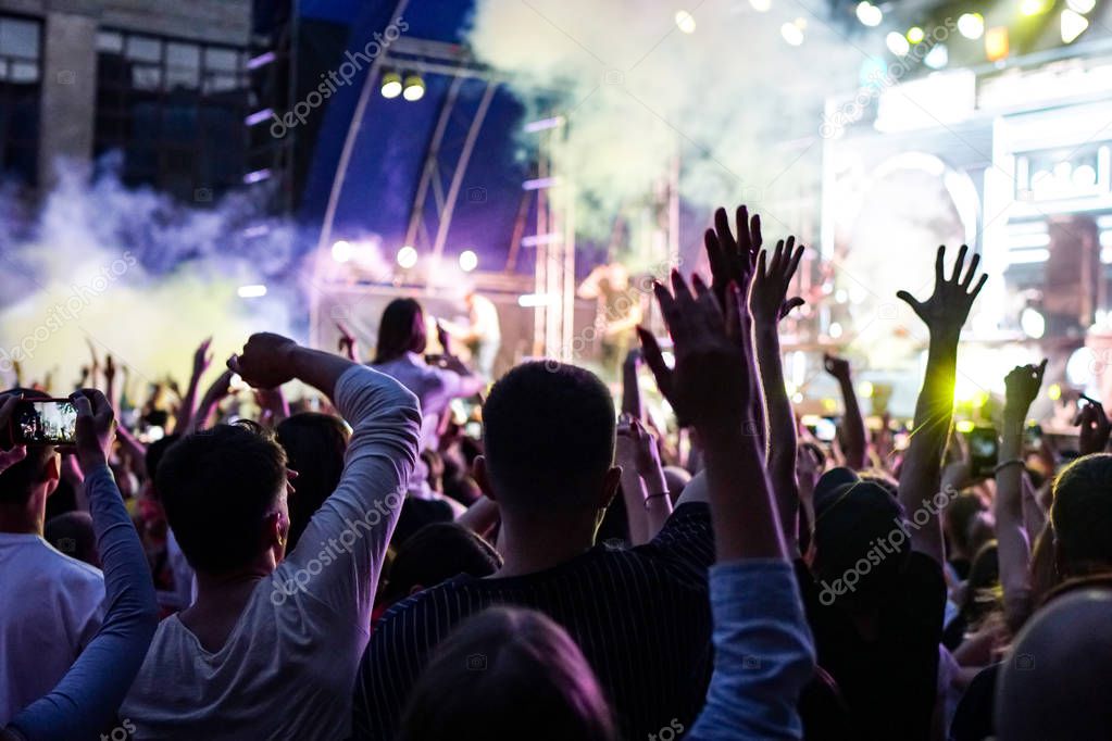 Concert crowd attending a concert, people silhouettes are visible, backlit by stage lights. Raised hands and smart phones are visible here and there.