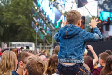 Çocuk bir açık hava rock müzik konserinde onlarla el ele tutuşarak ailesinin omuzlarında eğlenceli