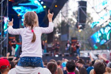 Çocuk bir açık hava rock müzik konserinde onlarla el ele tutuşarak ailesinin omuzlarında eğlenceli