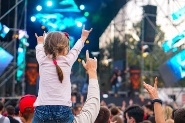 Çocuk bir açık hava rock müzik konserinde onlarla el ele tutuşarak ailesinin omuzlarında eğlenceli