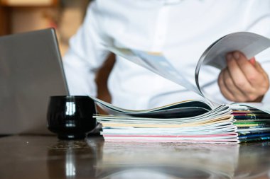 Yönetici dergilere ve dizüstü bilgisayarlara bakıyor, ön planda dergiler var.