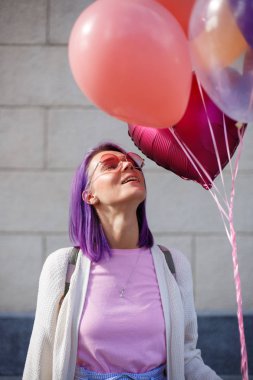 Balonlar yukarı bakıyor ve gülümseyen gözlük mor saçlı genç bayan